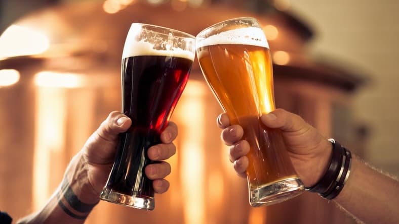 Image of hands toasting with glasses of craft beer in front of distillery vat.